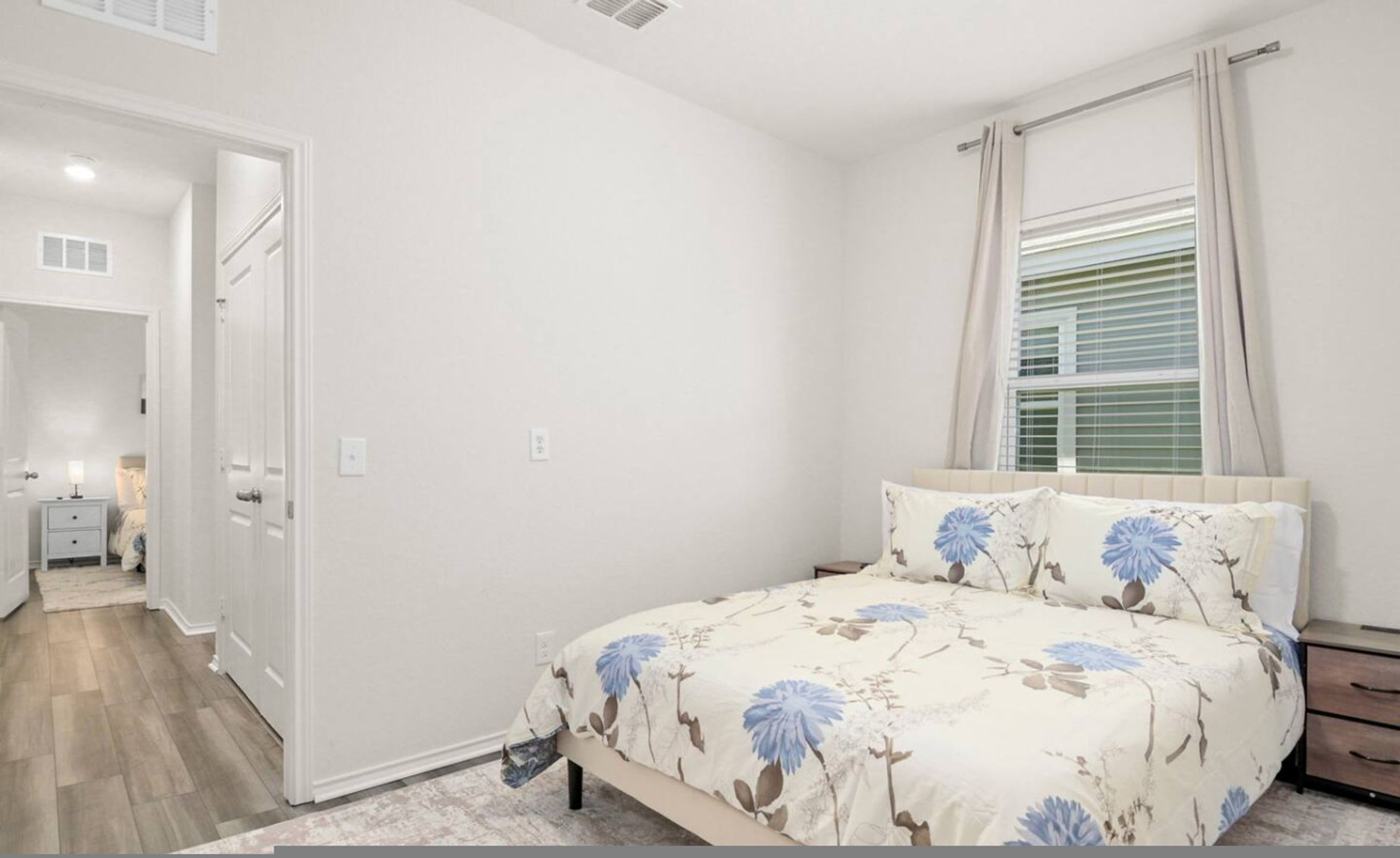 A bedroom with a bed, a nightstand, a window with blinds, and a door leading to another room.
