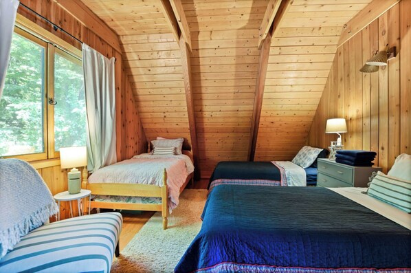 A cozy cabin bedroom with two beds, a window with curtains, a lamp, and a small table. - Lakefront A-Frame, Stunning Views, Private Dock (Merrimac)
