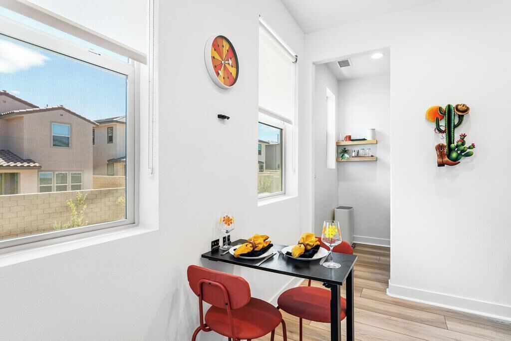 A dining area with a table set for two, a red chair, a clock on the wall, and a window overlooking a building.