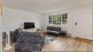 Un salon moderne avec un canapé gris, un téléviseur à écran plat, une table basse et une grande fenêtre donnant sur des arbres.