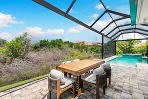A covered outdoor patio with a pool, wooden table, and chairs. - Tranquil Island Retreat. (Marco Island)