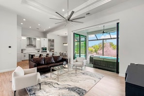 A spacious living area with a modern ceiling fan, a dark brown leather sofa, a glass coffee table, and a large window offering a view of a swimming pool and greenery. - Tranquil Island Retreat. (Marco Island)