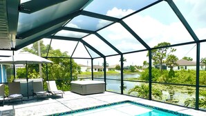 A covered outdoor swimming pool with lounge chairs and a hot tub, surrounded by a metal frame and glass walls, overlooking a body of water and greenery.