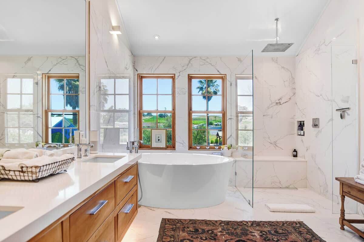 Ein modernes Badezimmer mit einer großen Badewanne, einer Dusche und einem Waschbecken. Große Fenster bieten Blick ins Grüne.