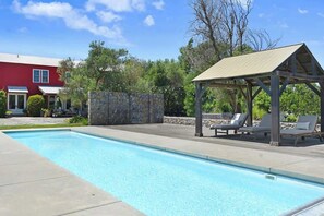 Une piscine avec des chaises longues et un kiosque, le tout entouré d’un mur de pierre et d’un bâtiment rouge.
