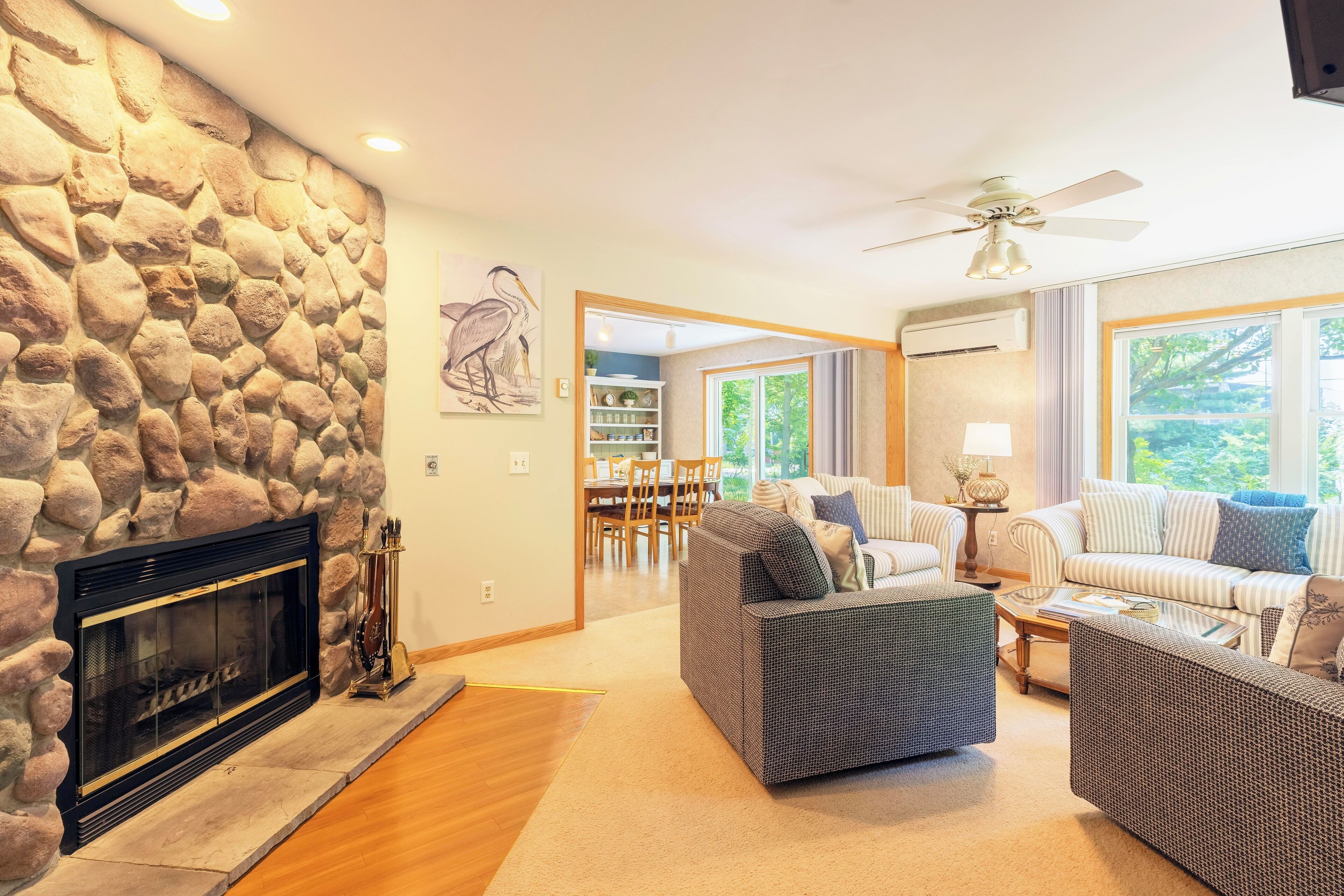 A living room with a stone fireplace, a ceiling fan, and a view of the outdoors through large windows.