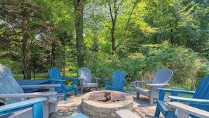 A circular stone fire pit surrounded by blue Adirondack chairs in a forested area.