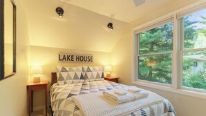 A bedroom with a bed, two bedside lamps, a window with a view of trees, and a sign that reads "LAKE HOUSE".