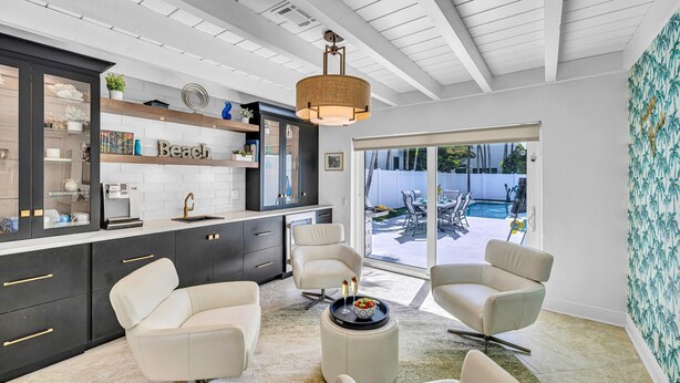 A modern kitchen with dark cabinets, a white countertop, and a large window. There are two white chairs and a round ottoman with a tray of drinks. - Steps to Beach / Heated Pool / Kiwi Key / Residences (Fort Lauderdale)