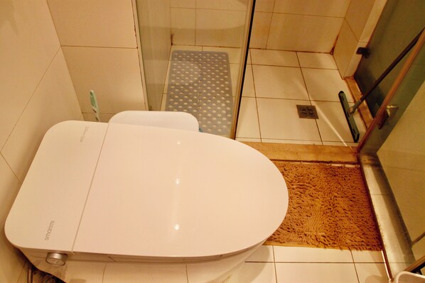 Un baño con inodoro blanco, zona de ducha y un tapete de baño marrón.
