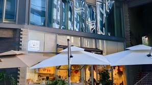 Ein modernes Gebäude mit großen Fenstern und einem Café namens Buddy Café. Es gibt Außenbereiche zum Sitzen mit Sonnenschirmen und Blumentöpfen.