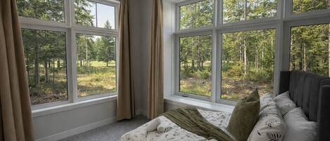 A bedroom with a large window offering a forest view, a bed with patterned bedding, and a headboard.
