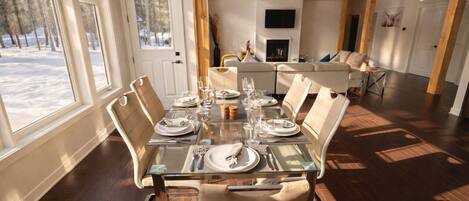 Un comedor con una mesa de vidrio dispuesta para seis personas, rodeada de sillas color beige. Se observa una chimenea y un televisor al fondo.