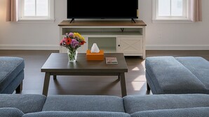 Ett vardagsrum med en platt-TV på ett trästativ, två fönster med gardiner, en tavla ovanför TV:n, ett soffbord med en vas med blommor och en soffa.