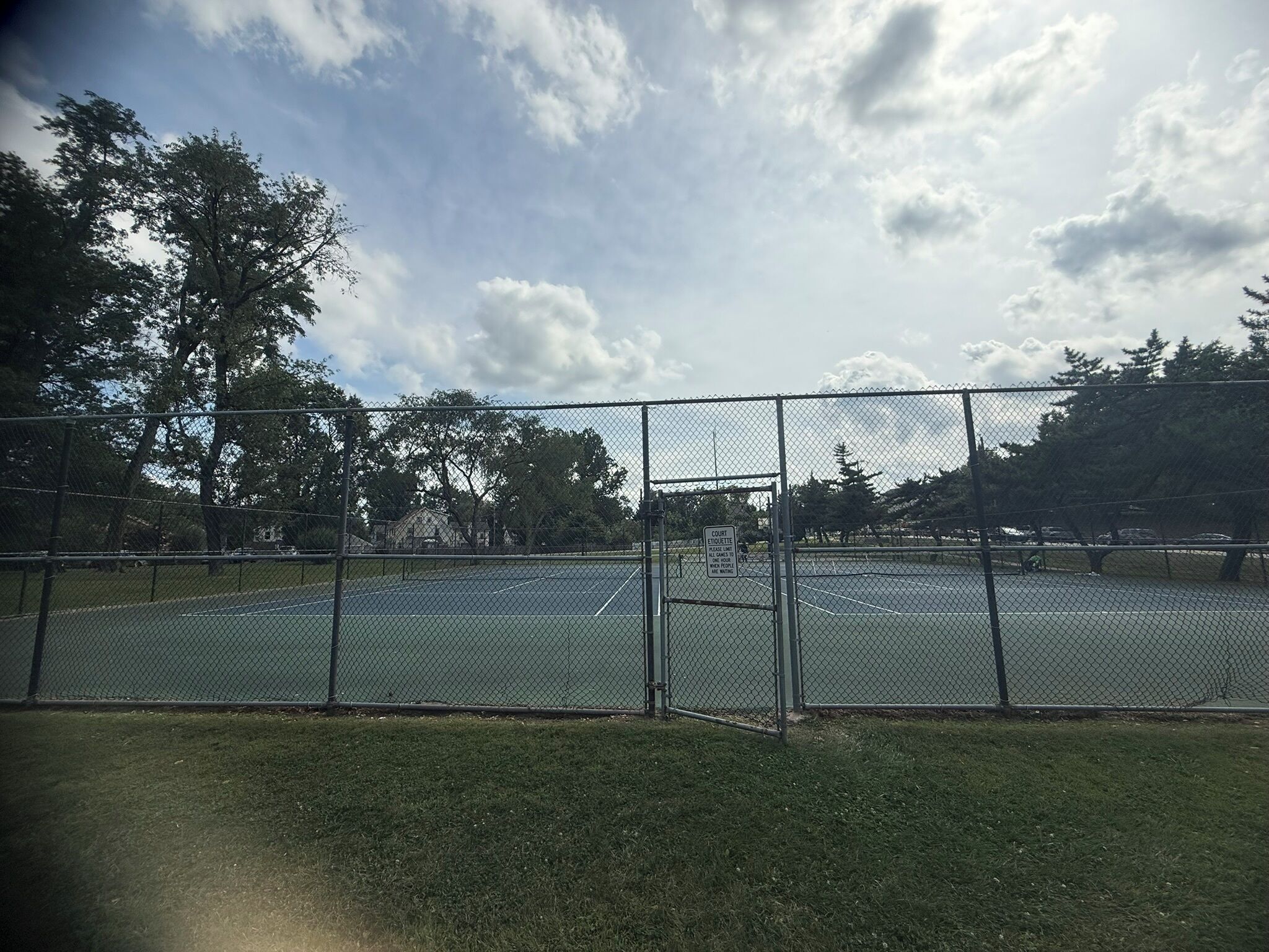 A fenced tennis court with a grassy area in front, surrounded by trees and a cloudy sky.