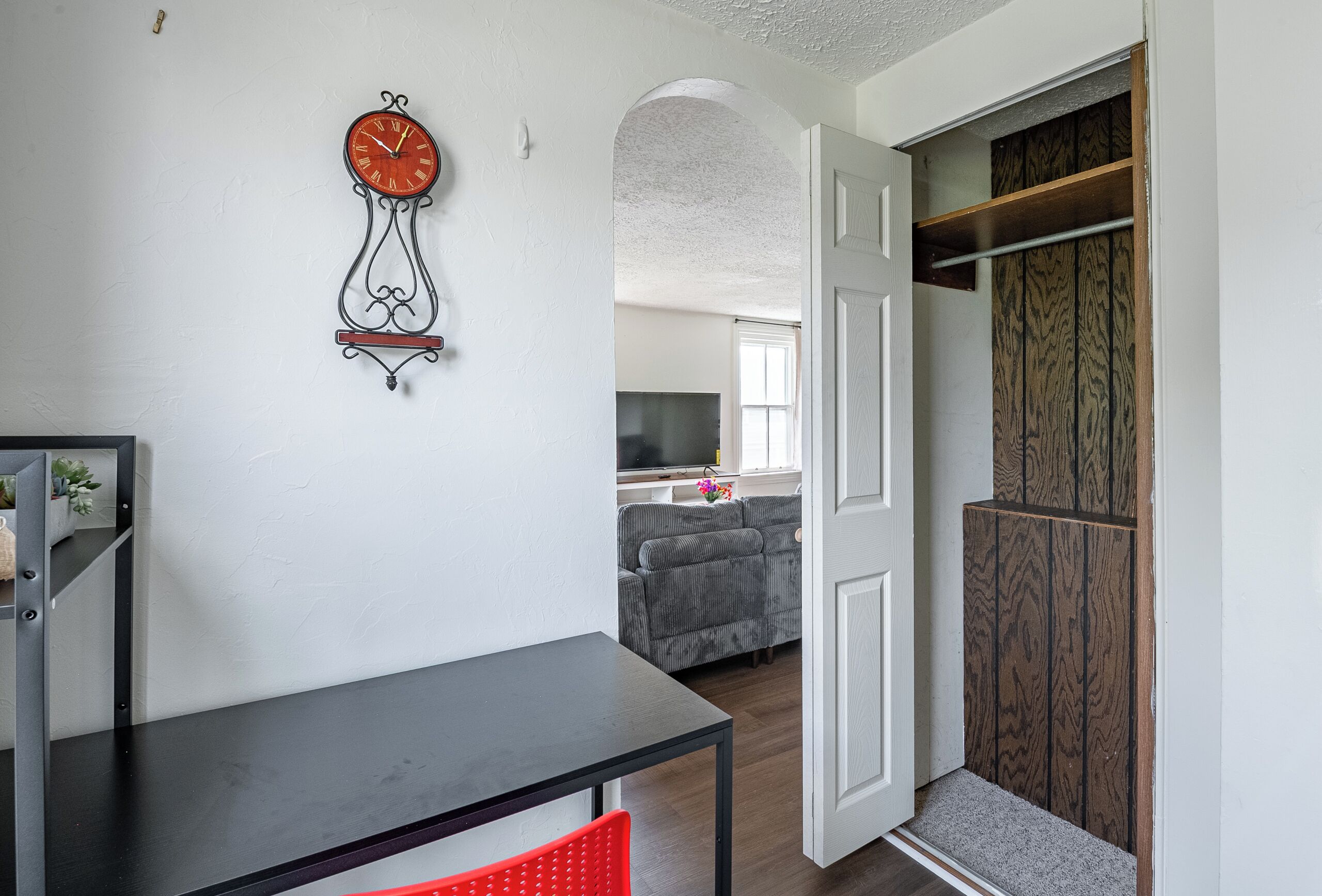 A room with a black table, a red chair, a clock on the wall, and a wooden wardrobe.