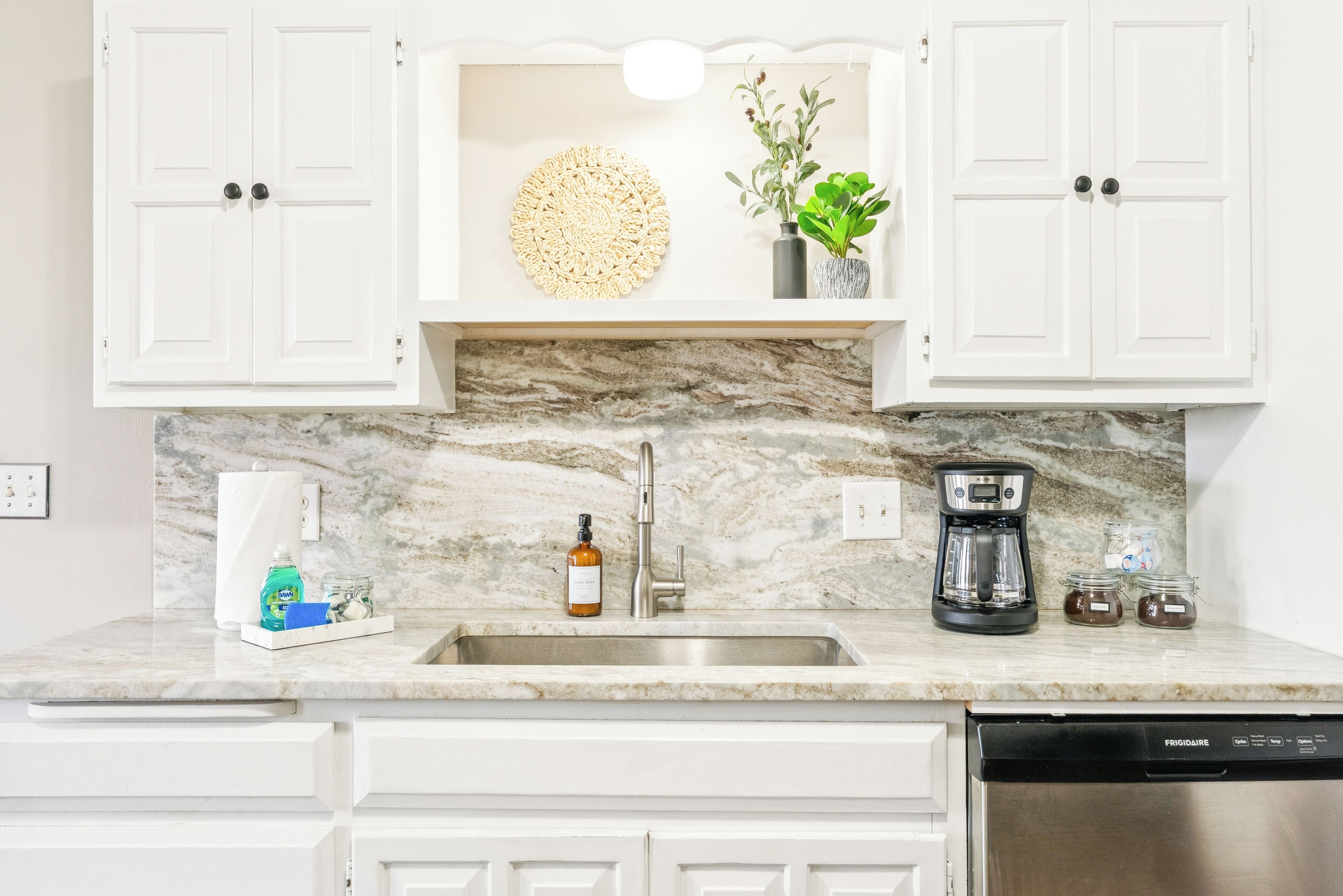 A kitchen with white cabinets, a marble backsplash, a coffee maker, a dish soap bottle, and a hand soap bottle.