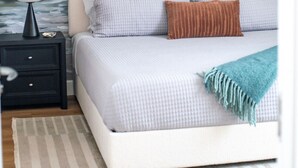 Un dormitorio con cama, almohadas, una mesita de noche, una lámpara y un mural en la pared que muestra una puesta de sol sobre el océano.