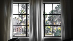 A room with two large windows, white curtains, and a view of palm trees and buildings outside. - Venus D | Futuristic 2BR + Pool & Near Beach (South Padre Island)