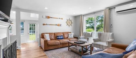 Uma sala de estar espaçosa com um sofá de couro marrom, uma poltrona listrada e uma mesa de centro. Há duas janelas com cortinas, um ar condicionado fixado na parede e uma porta que dá para o exterior.
