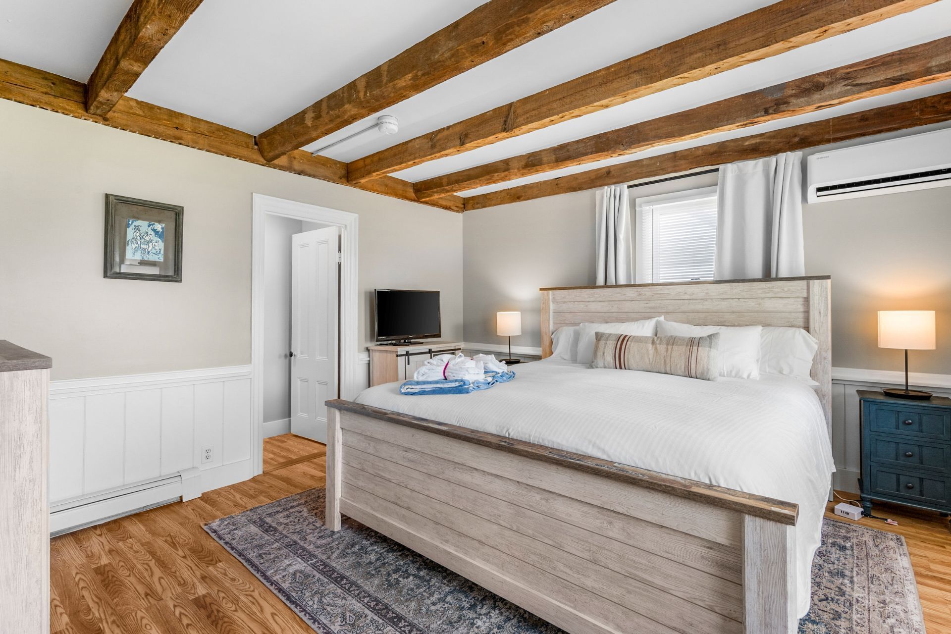 A bedroom with a wooden bed, white walls, a wooden ceiling, a television, and a window with curtains.