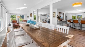 Un'ampia zona pranzo con un lungo tavolo in legno, sedie bianche e una cucina sullo sfondo.