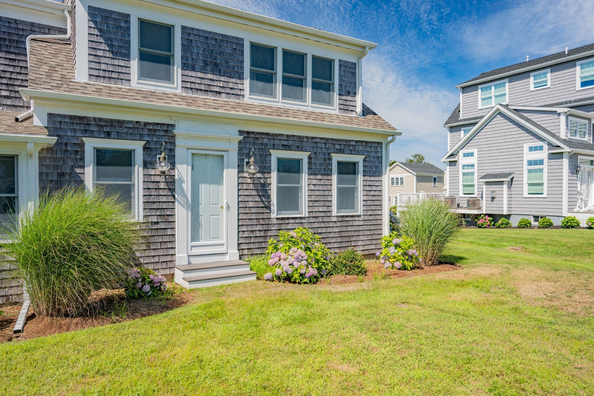 A two-story house with a front porch, a small garden with hydrangeas, and a clear blue sky.