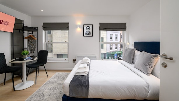 Een moderne hotelkamer met een bed, nachtkastje, stoel, bureau en uitzicht op de stad door grote ramen.