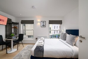 Ein modernes Hotelzimmer mit einem Bett, einem Nachttisch und einem Schreibtisch mit Stuhl. Es gibt zwei Fenster, einen an der Wand befestigten Fernseher und ein gerahmtes Bild.