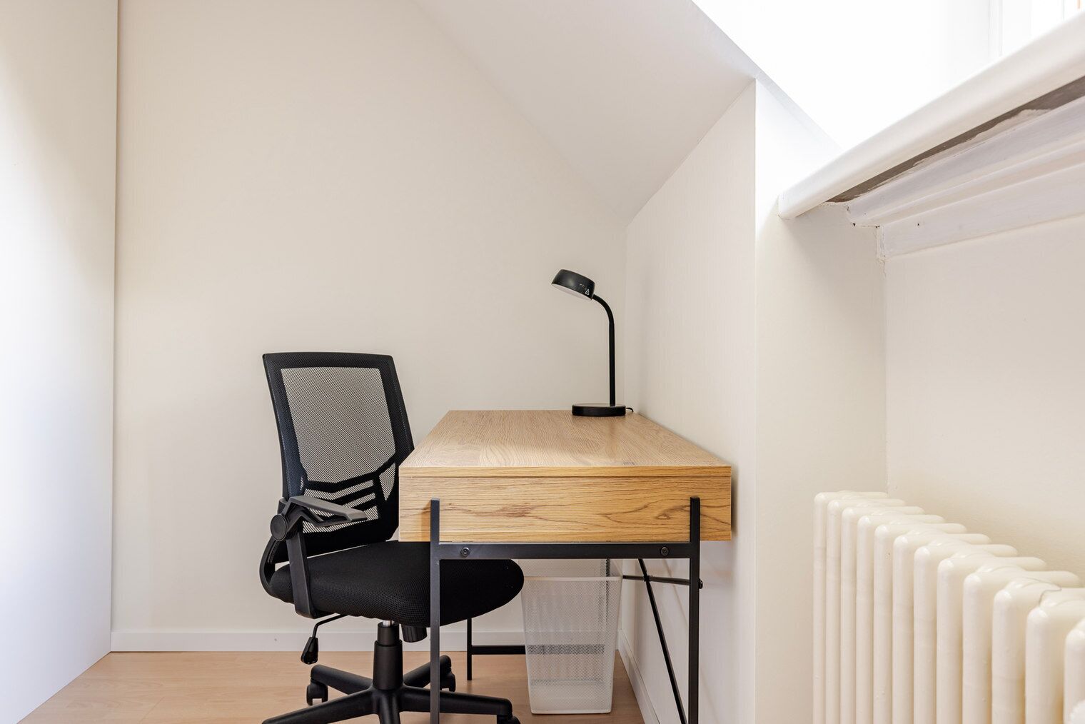 A small, well-lit room with a wooden desk, a black office chair, a black desk lamp, and a white radiator.