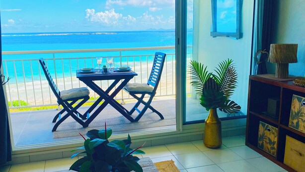 A room with a view of the ocean, a dining table set with plates and glasses, two chairs, a potted plant, and a wooden cabinet with decorative items.