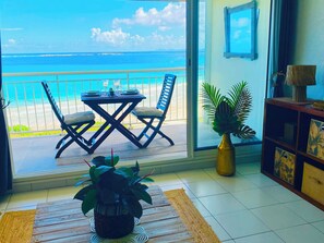 A room with a view of the ocean, a dining table set with plates and glasses, two chairs, a potted plant, and a wooden cabinet with decorative items.