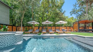 Une piscine dont les bords sont recouverts de carreaux, entourée de chaises longues et de parasols, avec une terrasse en bois et une clôture en bois en arrière-plan.