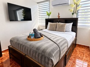 Habitación de hotel con cama, televisor de pantalla plana, ventana con persianas y aire acondicionado instalado en la pared.