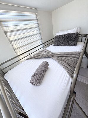 A single bed with a grey blanket and a rolled-up blanket, a grey pillow, and a window with blinds.