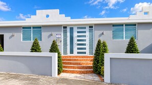 Un edificio moderno de dos pisos con fachada blanca, una escalera que lleva a la entrada y dos pequeños árboles a cada lado.
