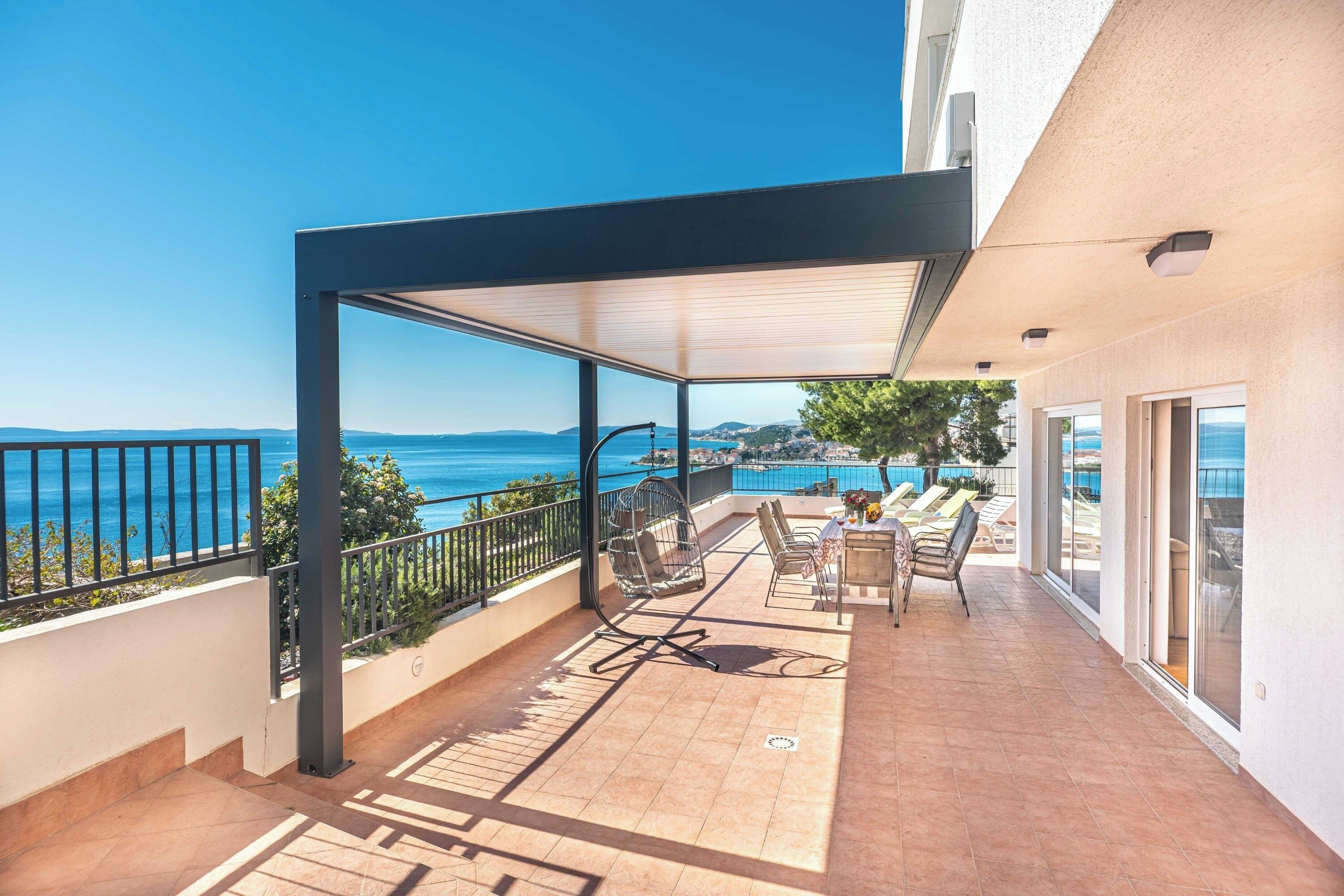 A covered outdoor area with a view of the sea, furnished with chairs and a table, and a railing along the edge.
