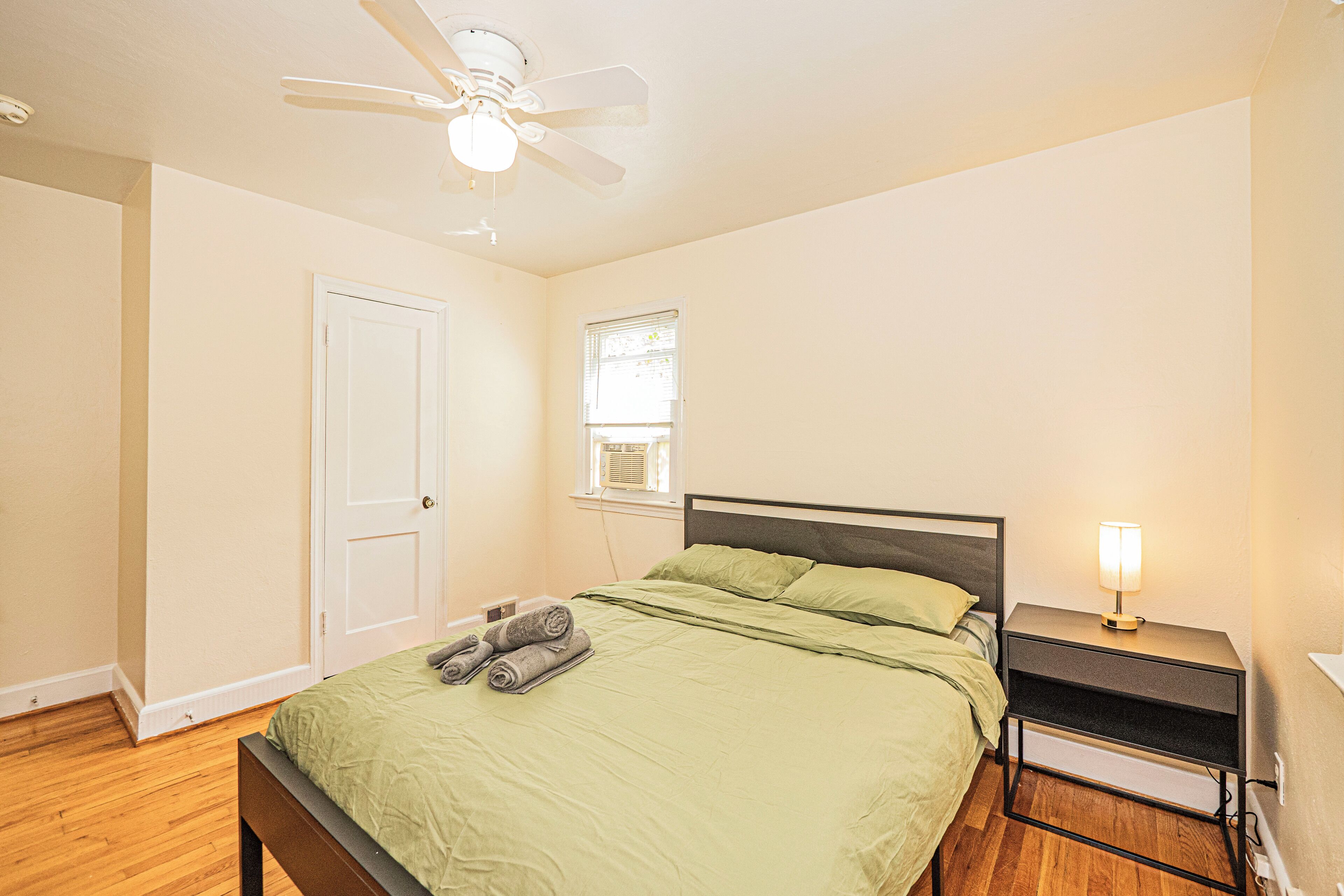 A bedroom with a bed, a nightstand with a lamp, a ceiling fan, a door, and a window.
