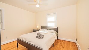 Un dormitorio con cama, ventilador de techo, mesita de noche, lámpara y ventana.