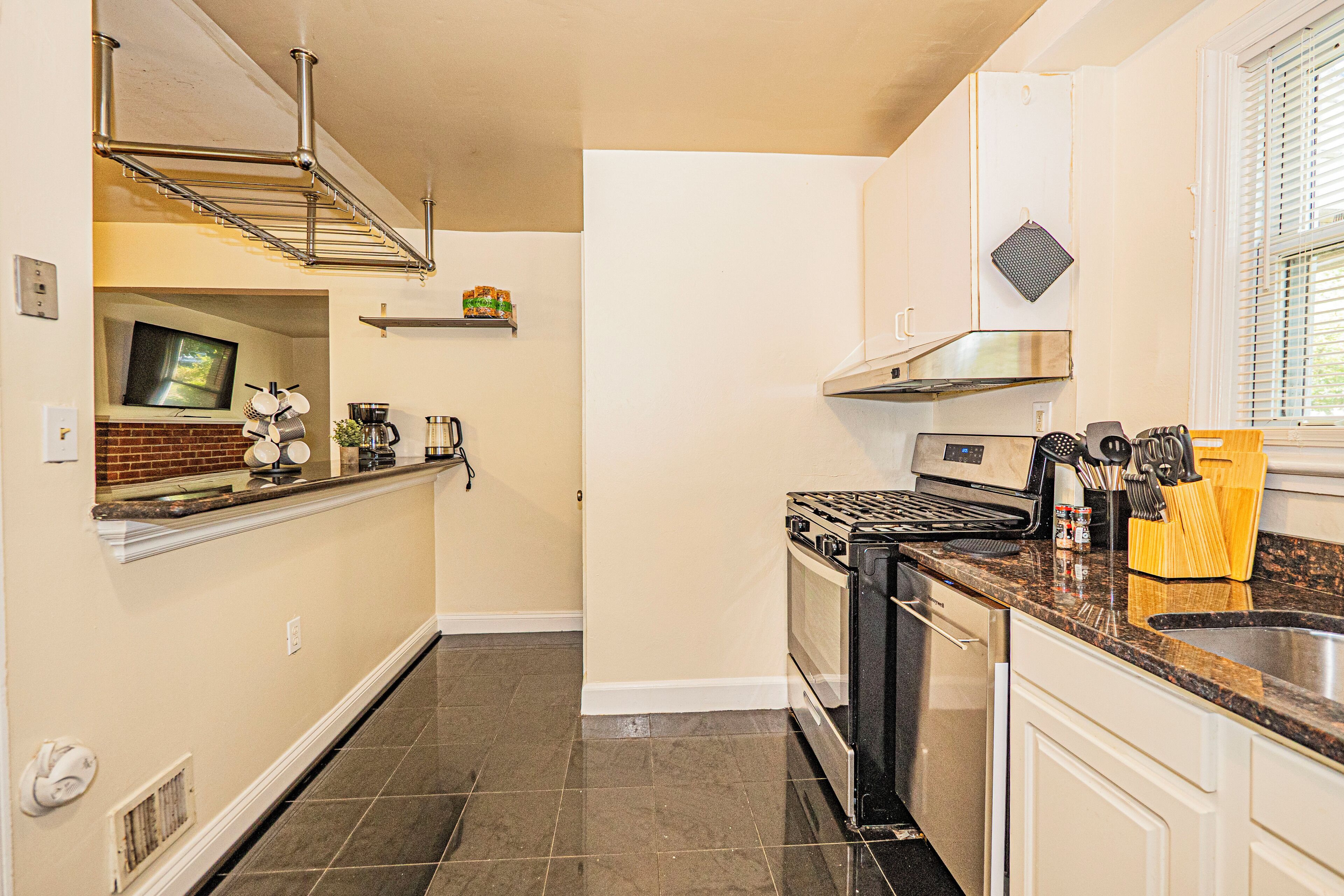 A kitchen with a stove, oven, and sink. There is a window with blinds, a shelf with kitchen items, and a vent on the wall.