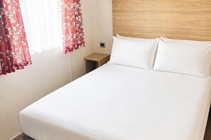 Een eenpersoonsbed met wit beddengoed in een kamer met een houten hoofdeinde, een klein houten nachtkastje en een raam met rode gordijnen met bloemenmotief.