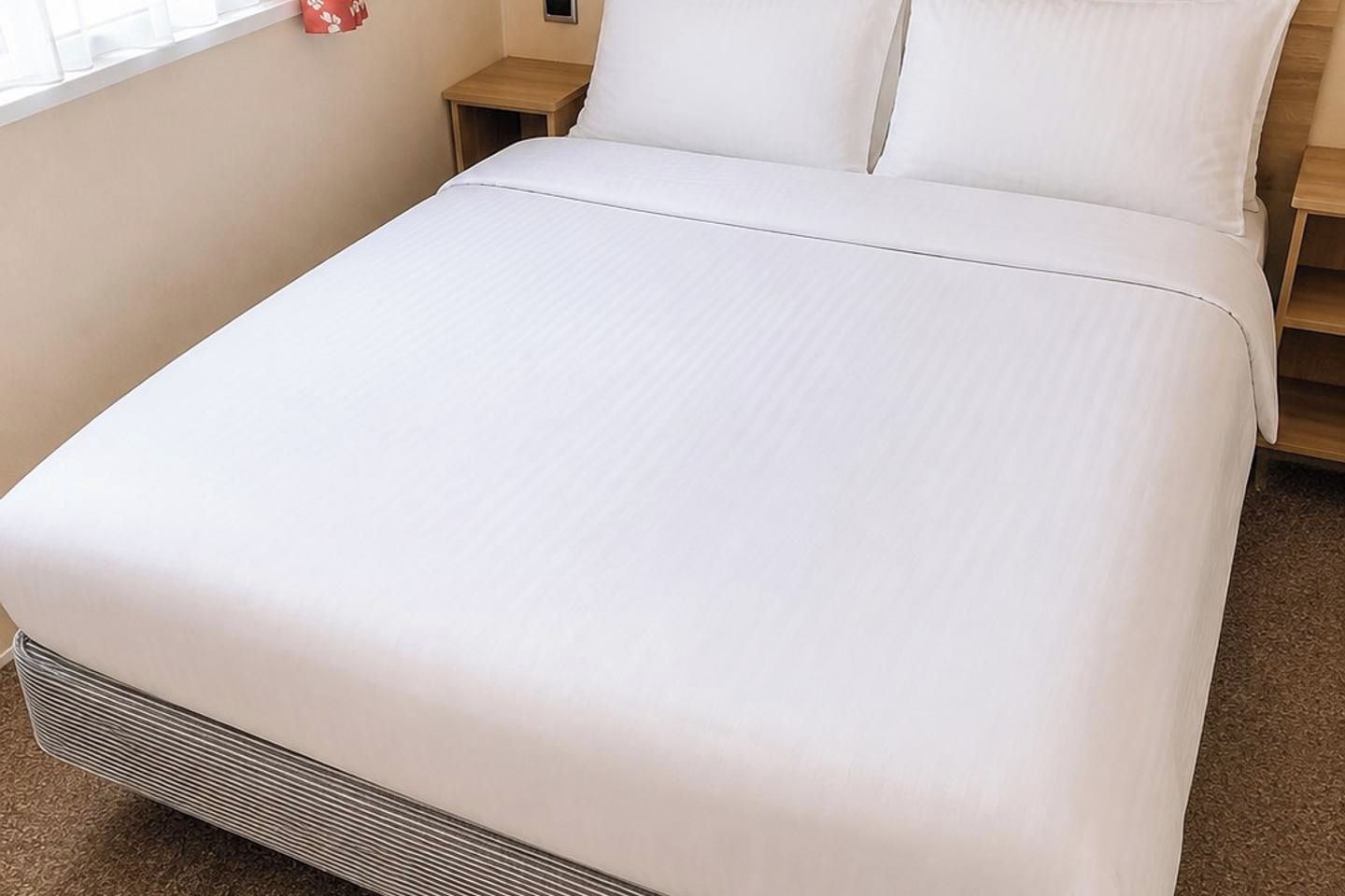 A neatly made bed with a white bedspread and pillows, a wooden nightstand with a lamp, and a small red object on the wall.