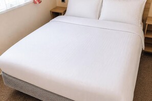 Una cama bien hecha con cubrecama y almohadas blancas, una mesita de noche de madera con una lámpara, y un objeto rojo pequeño en la pared.