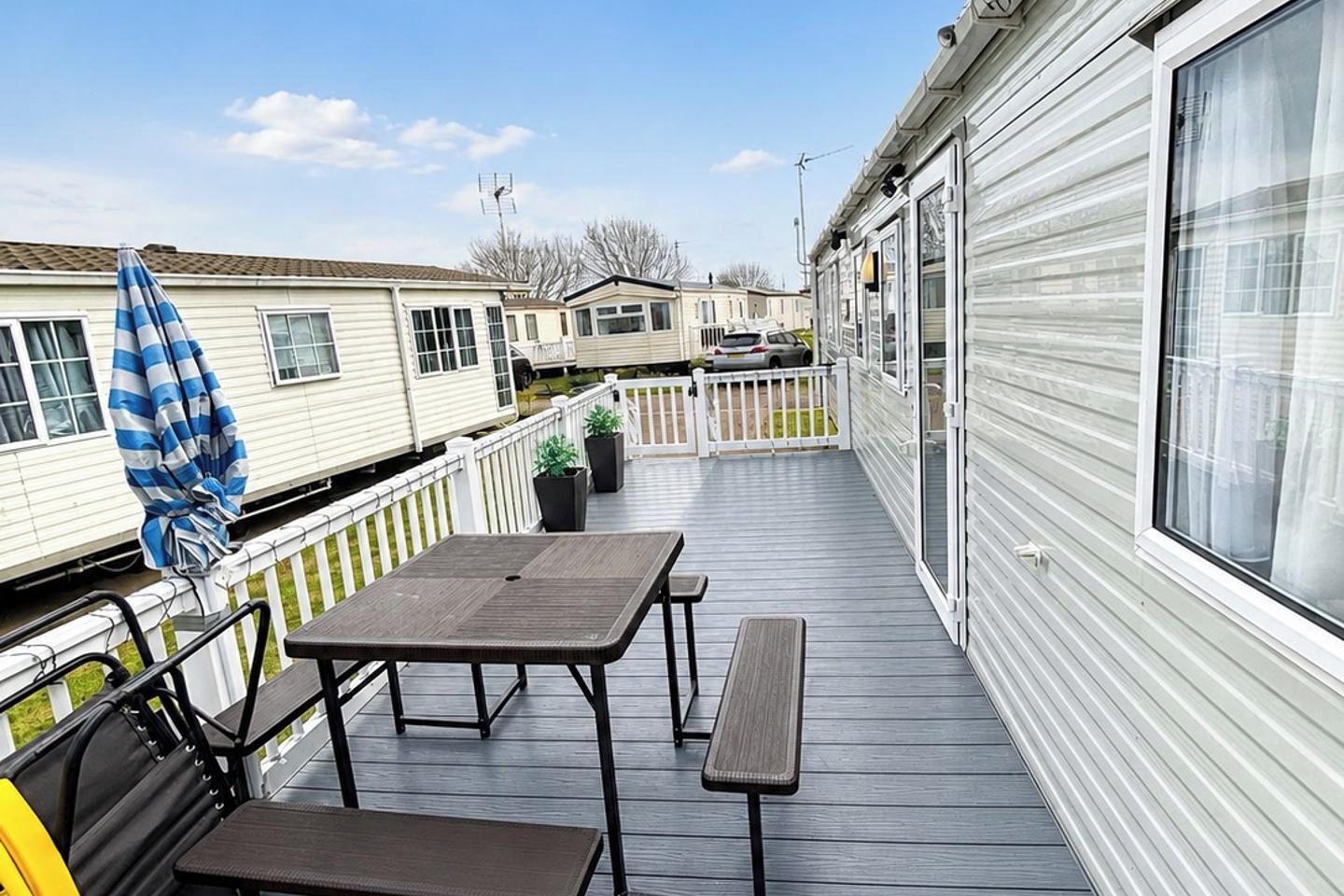A deck with a table and benches, surrounded by a white railing, with a view of other mobile homes in the background.
