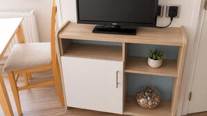 Une pièce avec un téléviseur posé sur un meuble, une armoire blanche avec un tiroir, une chaise et une étagère ornée d’une plante et d’un objet décoratif.