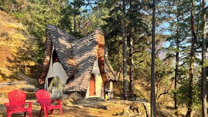 Une cabane rustique, dotée d’un toit à forte pente, est entourée d’une forêt. Deux chaises Adirondack rouges sont placées devant la cabane.