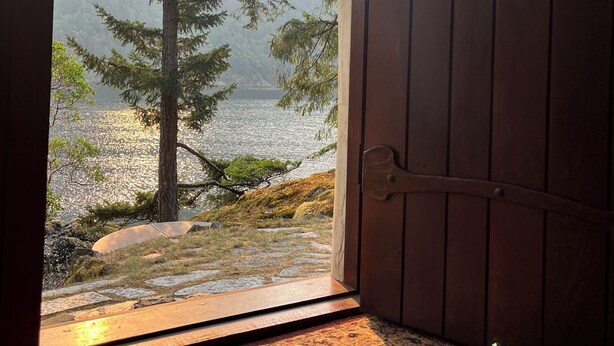Une porte en bois ouverte, donnant sur un paysage où l’on aperçoit un lac, entouré d’arbres et de montagnes.