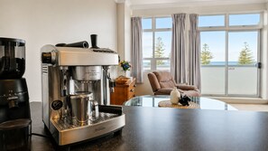 A modern living room with a coffee machine, a glass table, a chair, and a view of the outdoors.