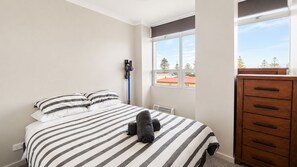 A bedroom with a bed, a dresser, a window with a view of trees and buildings, and a wall-mounted heater.