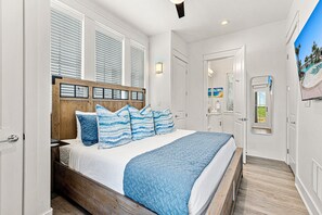 A bedroom with a wooden headboard, a large bed with blue bedding, a window with blinds, and a door leading to a bathroom. - Modern home w/ two master suites, golf course view - pool, beach, gym, & more (Port Aransas)